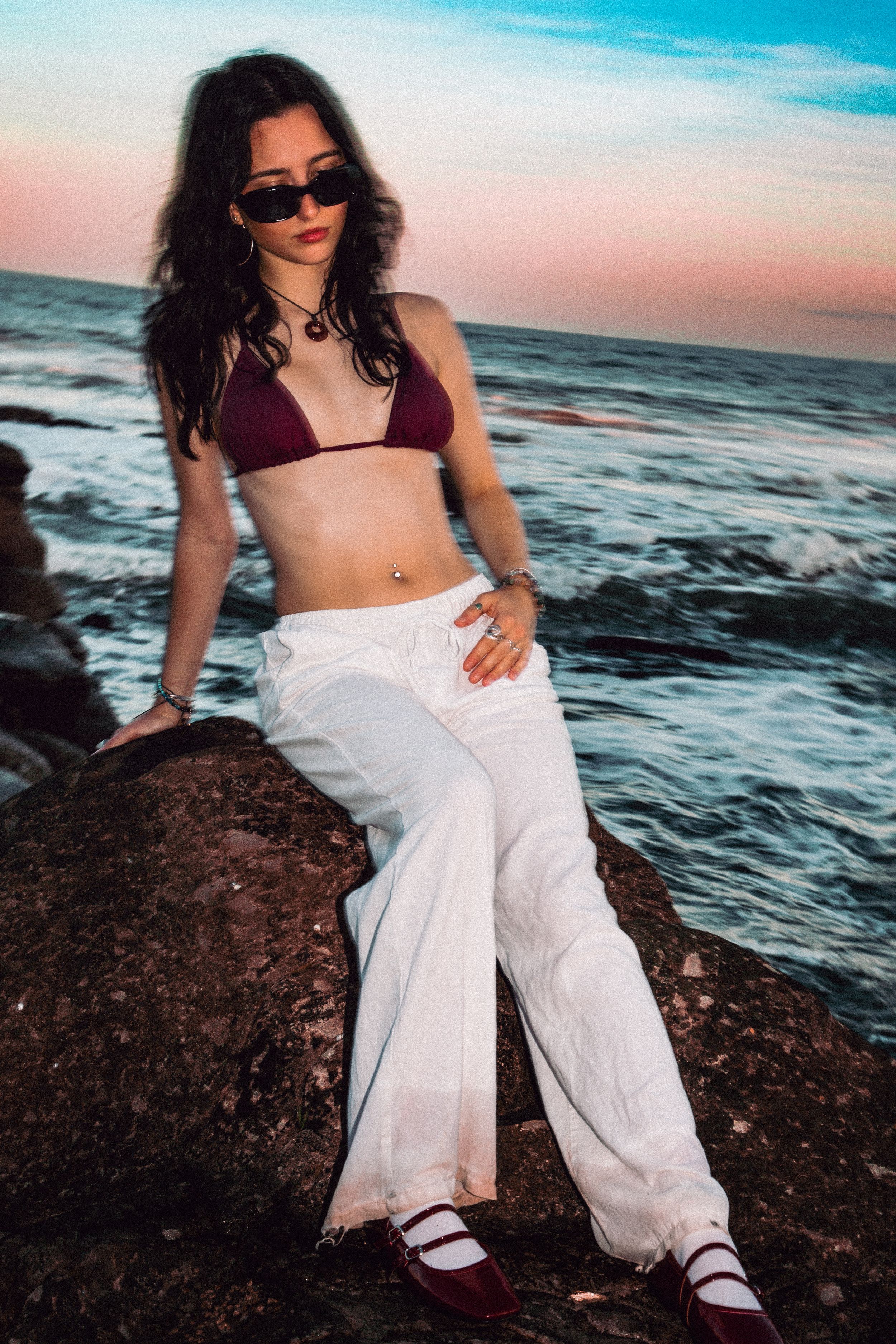 Casual beach fashion shoot featuring white pants and burgundy top on rocky shore.