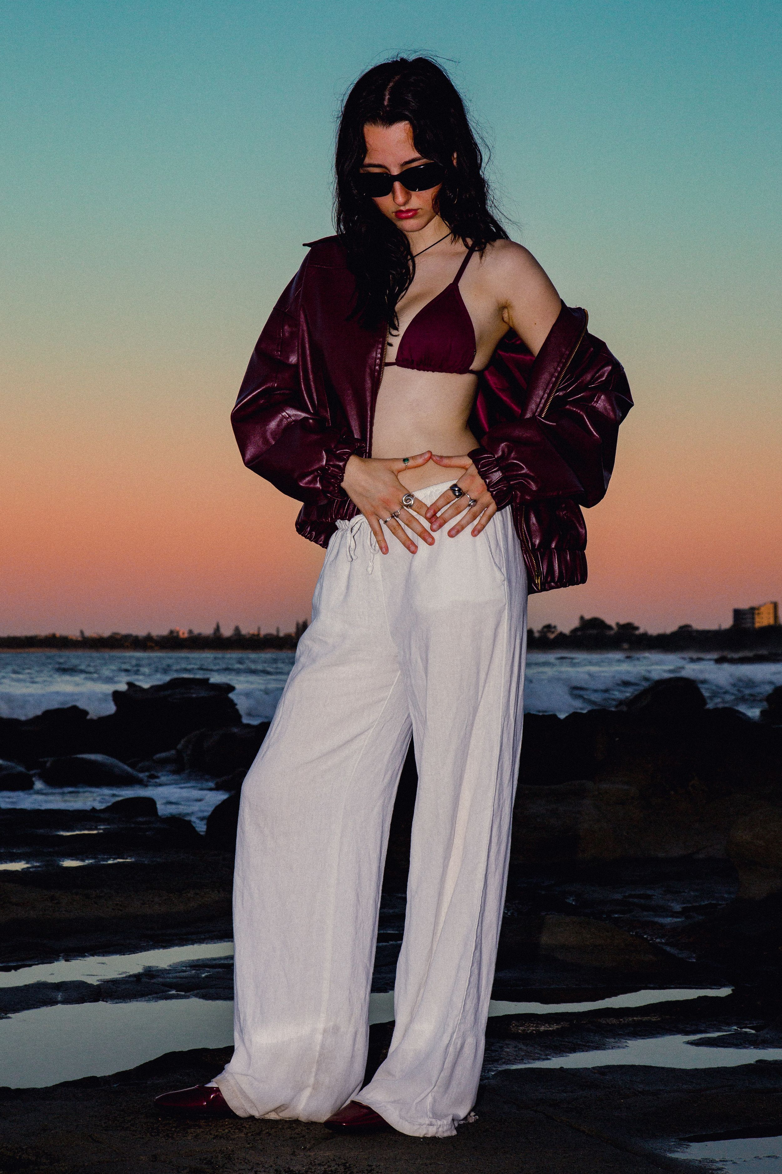 Fashion model poses at sunset wearing white wide-leg pants, burgundy crop top and jacket near rocky coastline.