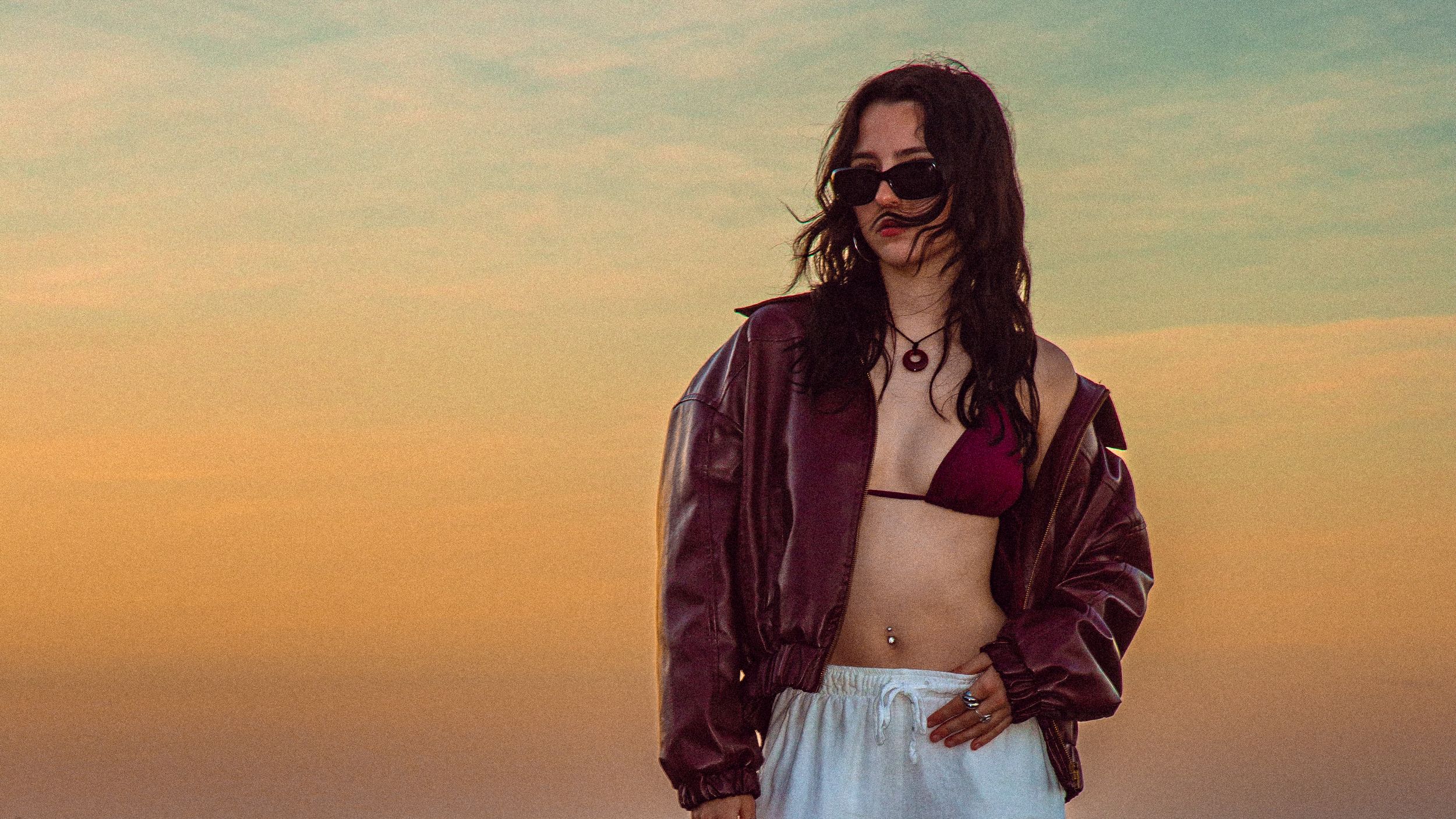 Sunset fashion portrait featuring burgundy jacket and crop top against orange sky.