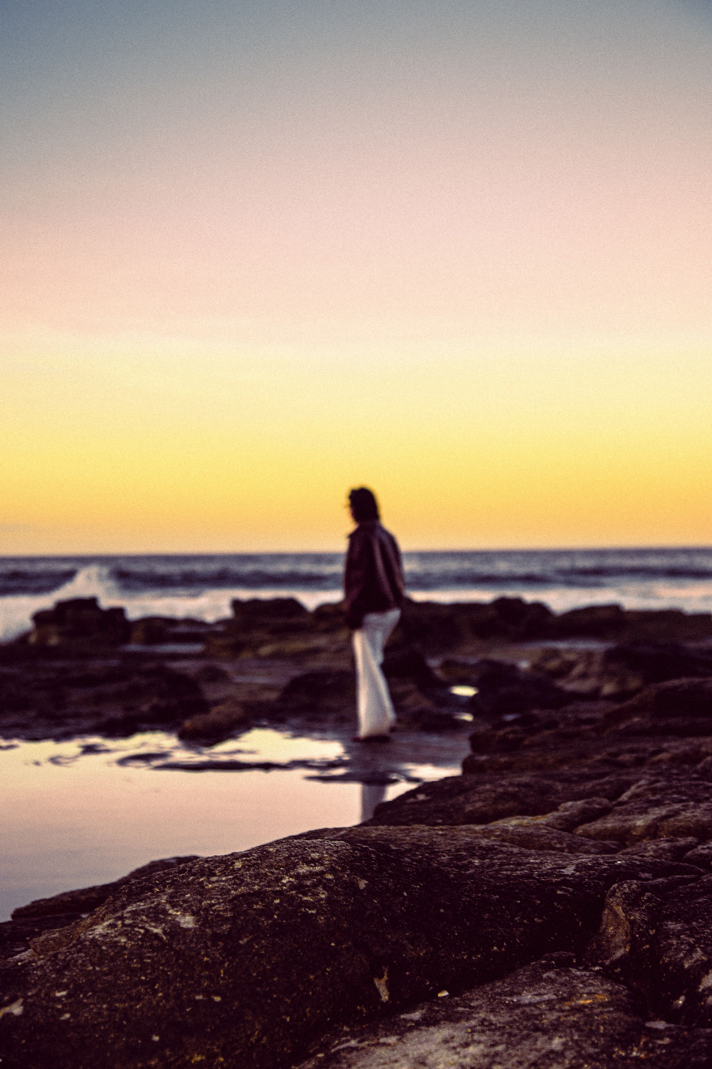 Silhouette walking on rocky coastline during golden sunset.