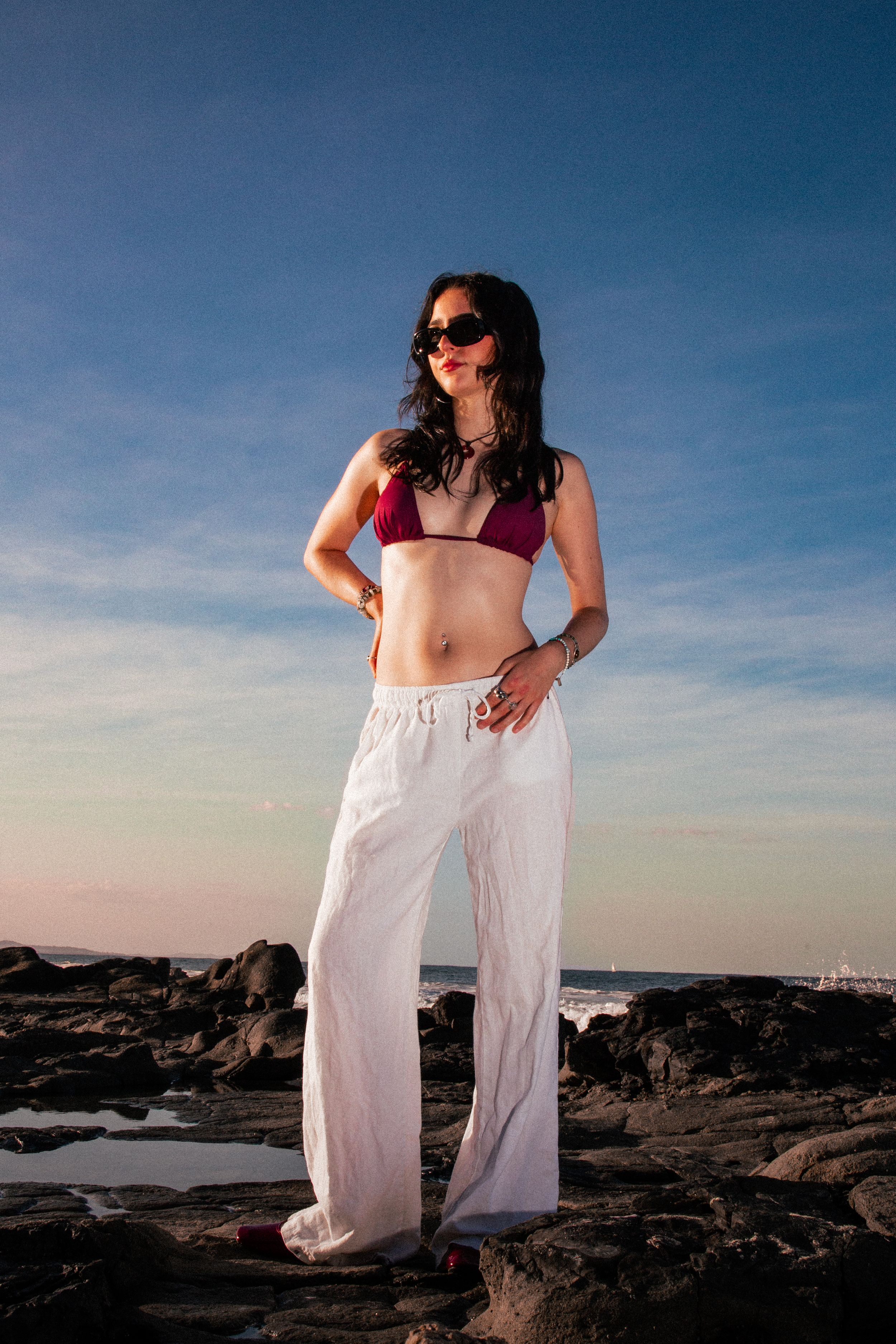 Fashion model in white pants and burgundy top poses dramatically on rocky beach at sunset.