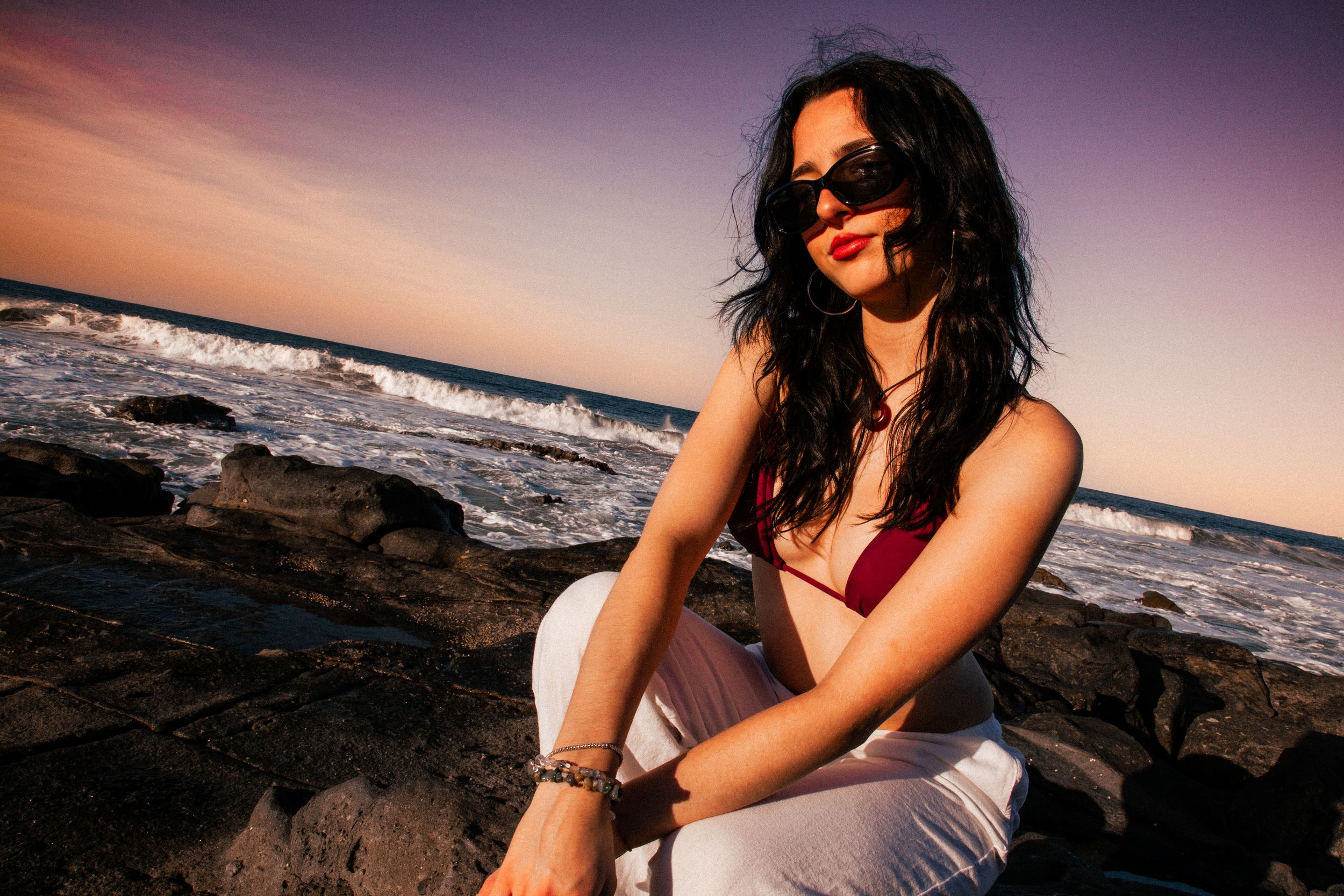 Beachside photoshoot featuring casual white pants and sunglasses against ocean waves at dusk.