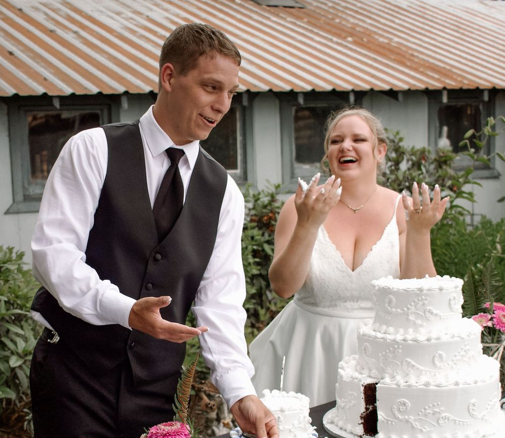 Smiling bride and groom stand beside a white tiered wedding cake at outdoor reception.