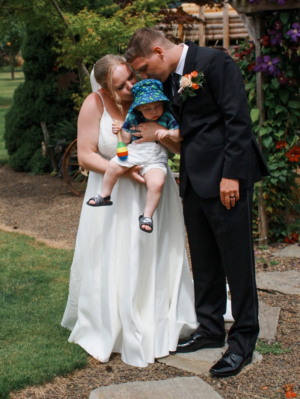 Bride and groom tenderly holding their baby during outdoor garden wedding ceremony.