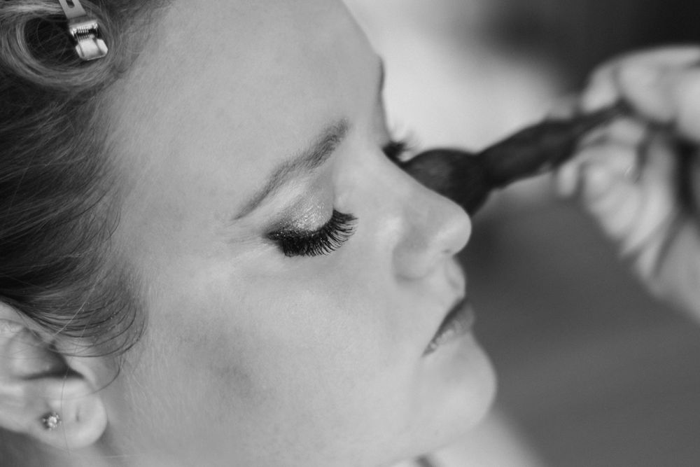 Black and white close-up of makeup artist applying eyeshadow to young woman with long lashes and ear piercing.