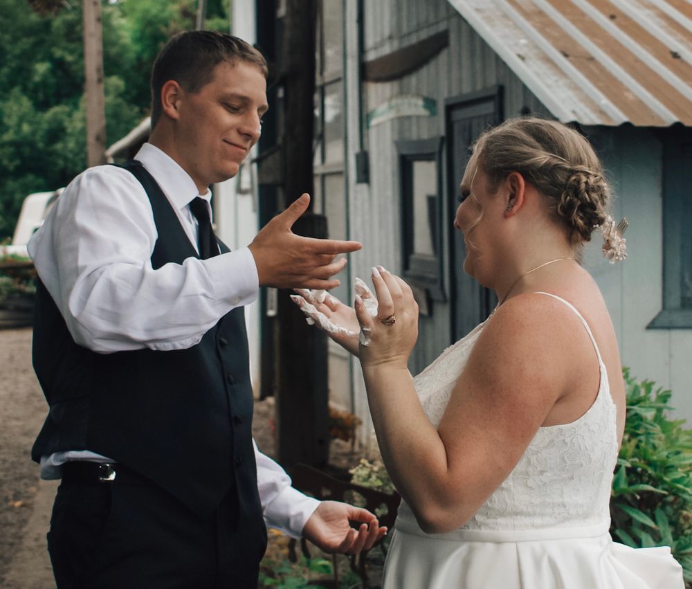 Bride and groom laugh together during playful cake smashing tradition at outdoor wedding.