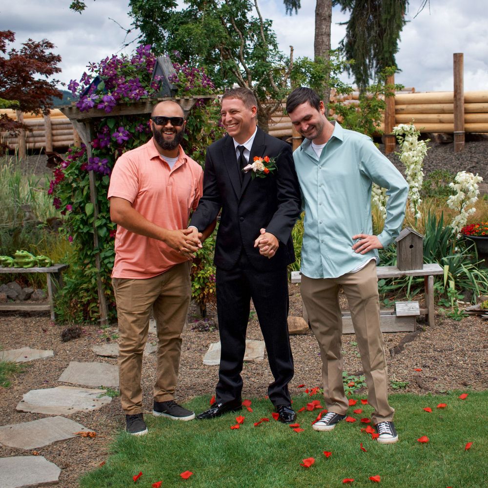 Groom in black suit poses with two friends in a colorful garden wedding venue.