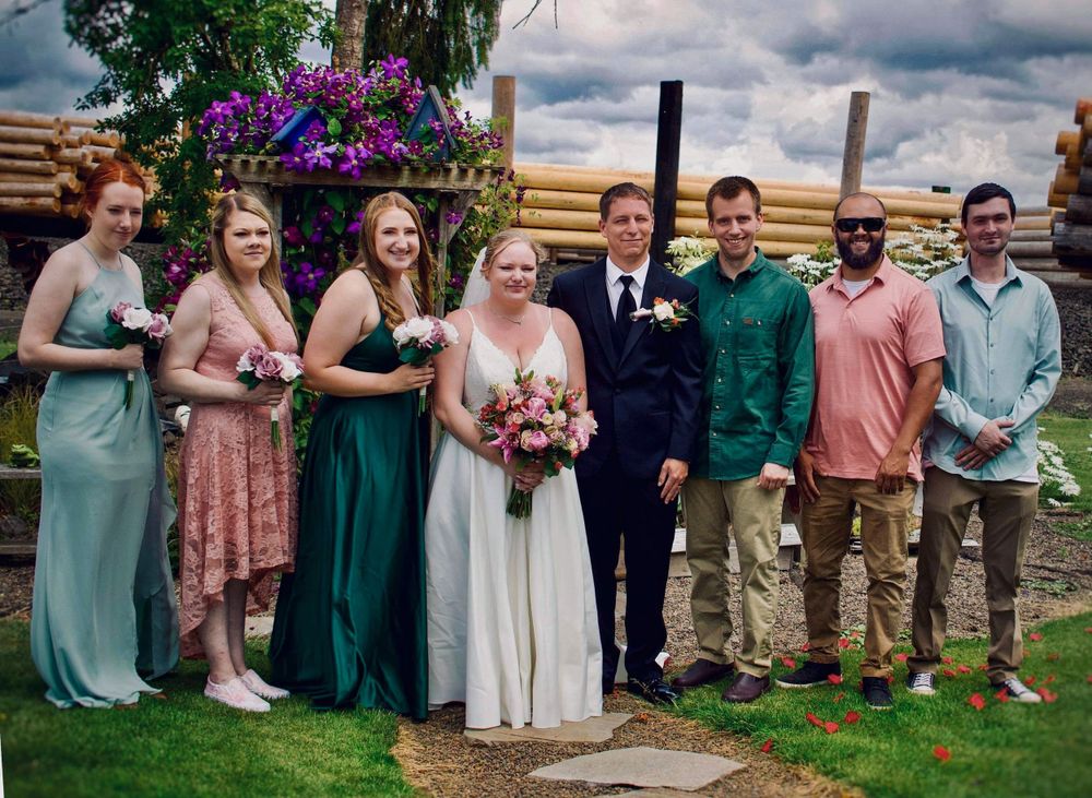 Wedding party poses together outdoors near a log cabin with colorful flowers in background.