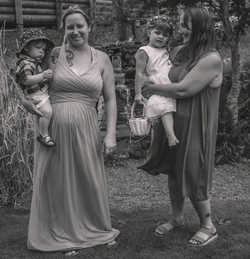 Two women holding toddlers pose outdoors in formal attire at a garden wedding celebration.