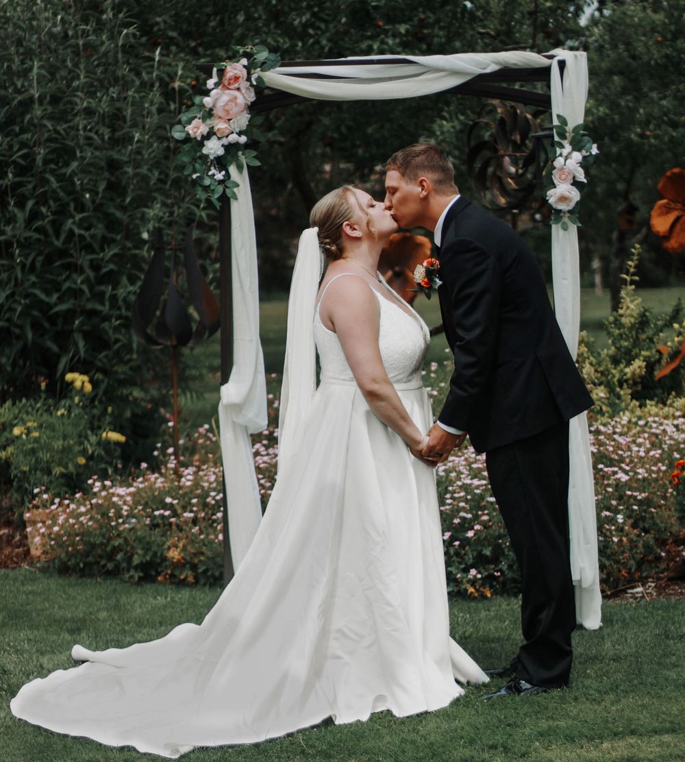 Bride and groom share a first kiss under a floral arch in a lush garden wedding ceremony.