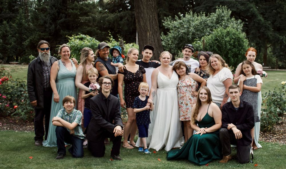 Large wedding party group photo taken outdoors surrounded by lush green trees and flowers.