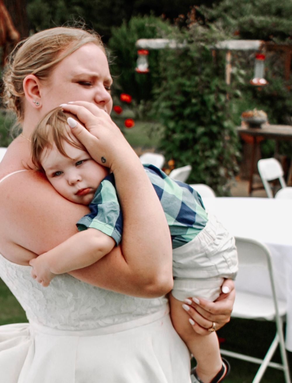 Bride in white wedding dress tenderly holds a young boy at an outdoor garden wedding reception.
