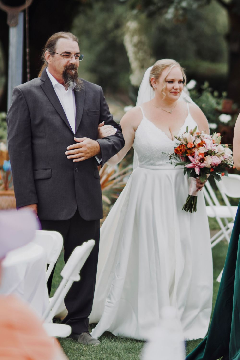 Bride walks down the aisle escorted by her father during outdoor garden wedding.