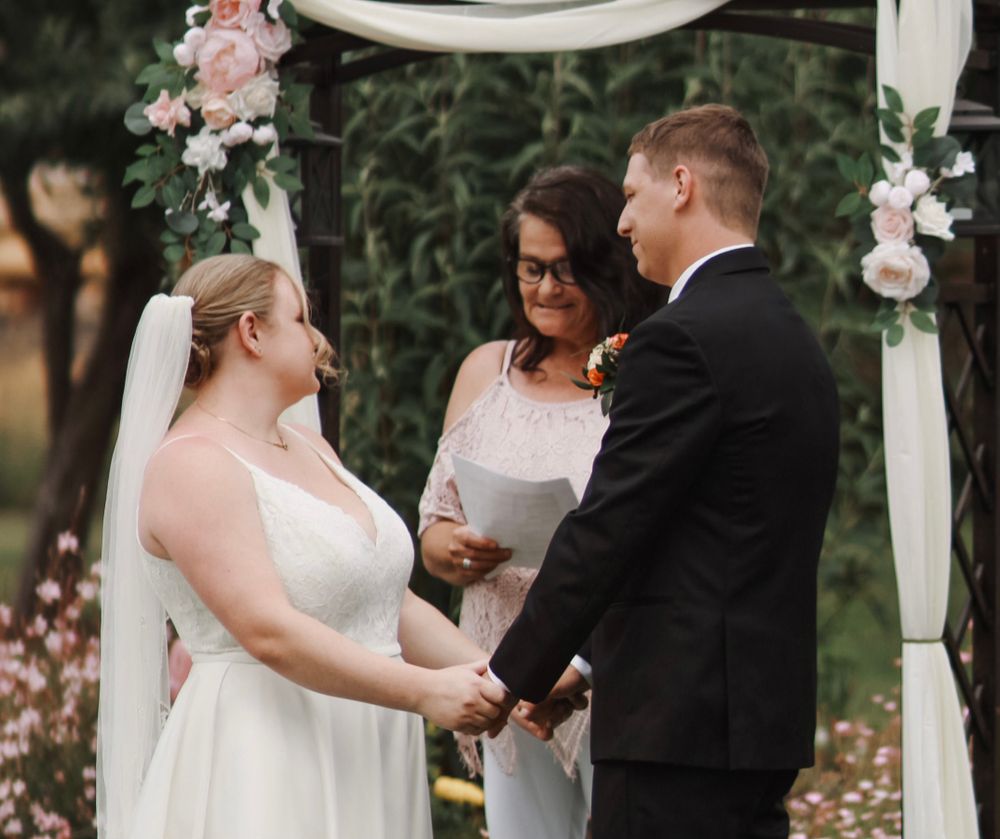 Bride and groom exchange vows before officiant under a white floral wedding arch.