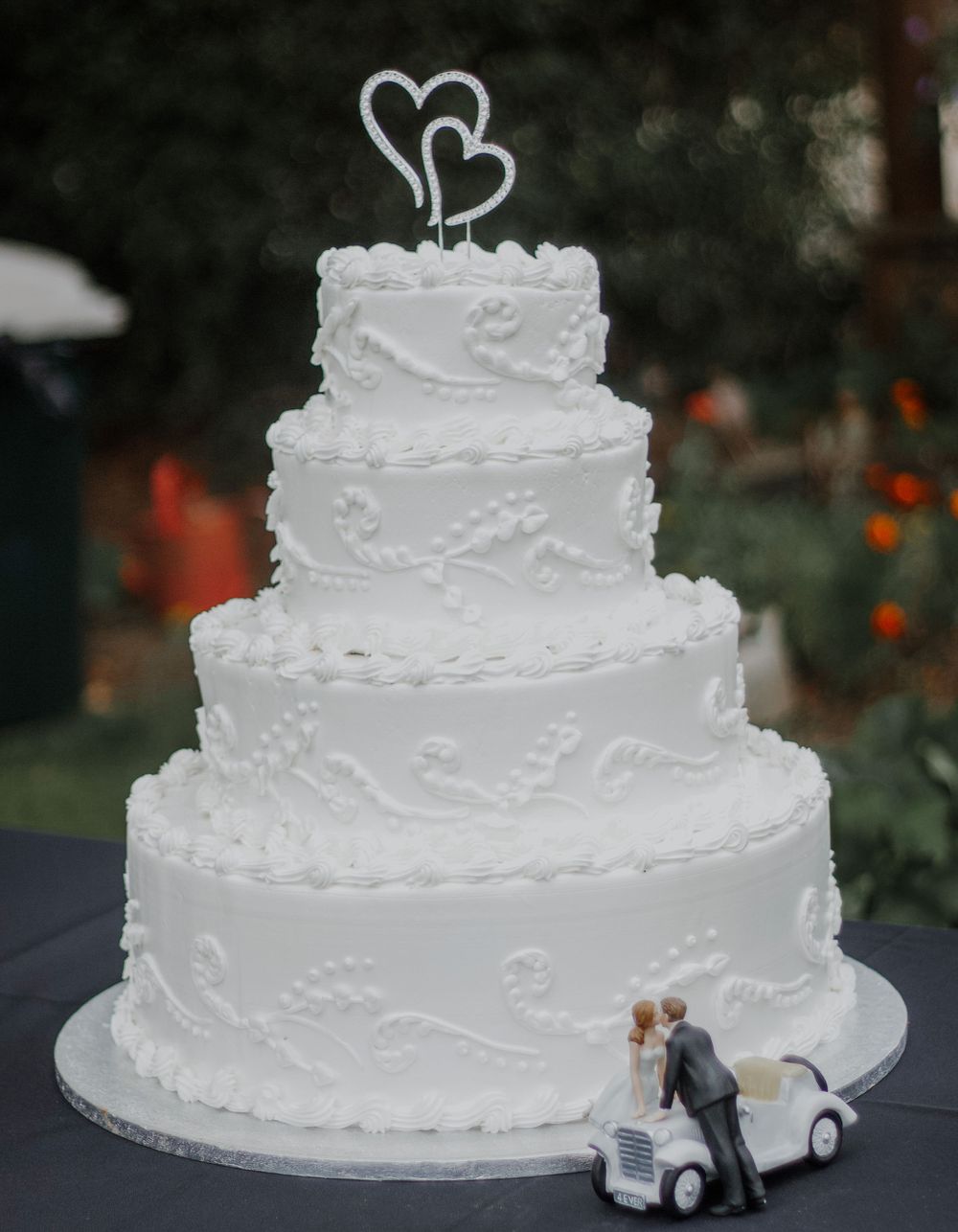 Four-tier white wedding cake with heart topper and bride and groom figurines at outdoor reception.