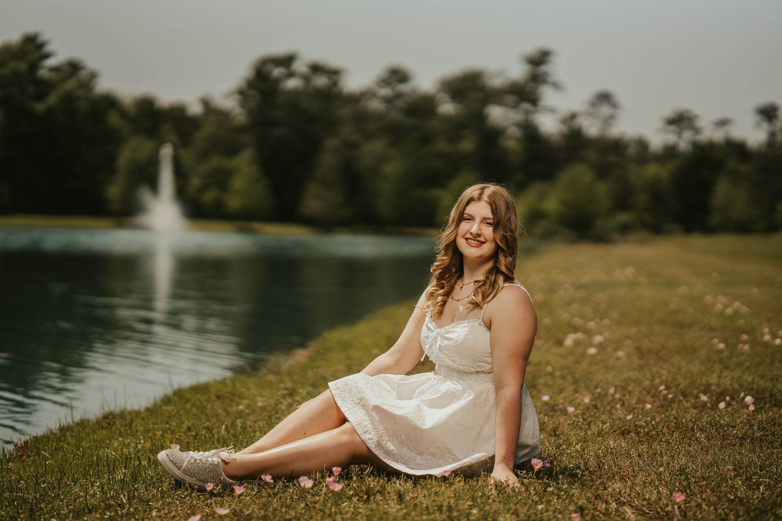 Woman in white dress sits by lake with fountain on summer evening.