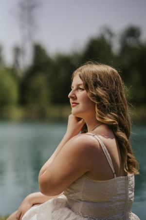 Portrait of a woman in a light dress sitting near a lake with soft lighting and trees in the background.