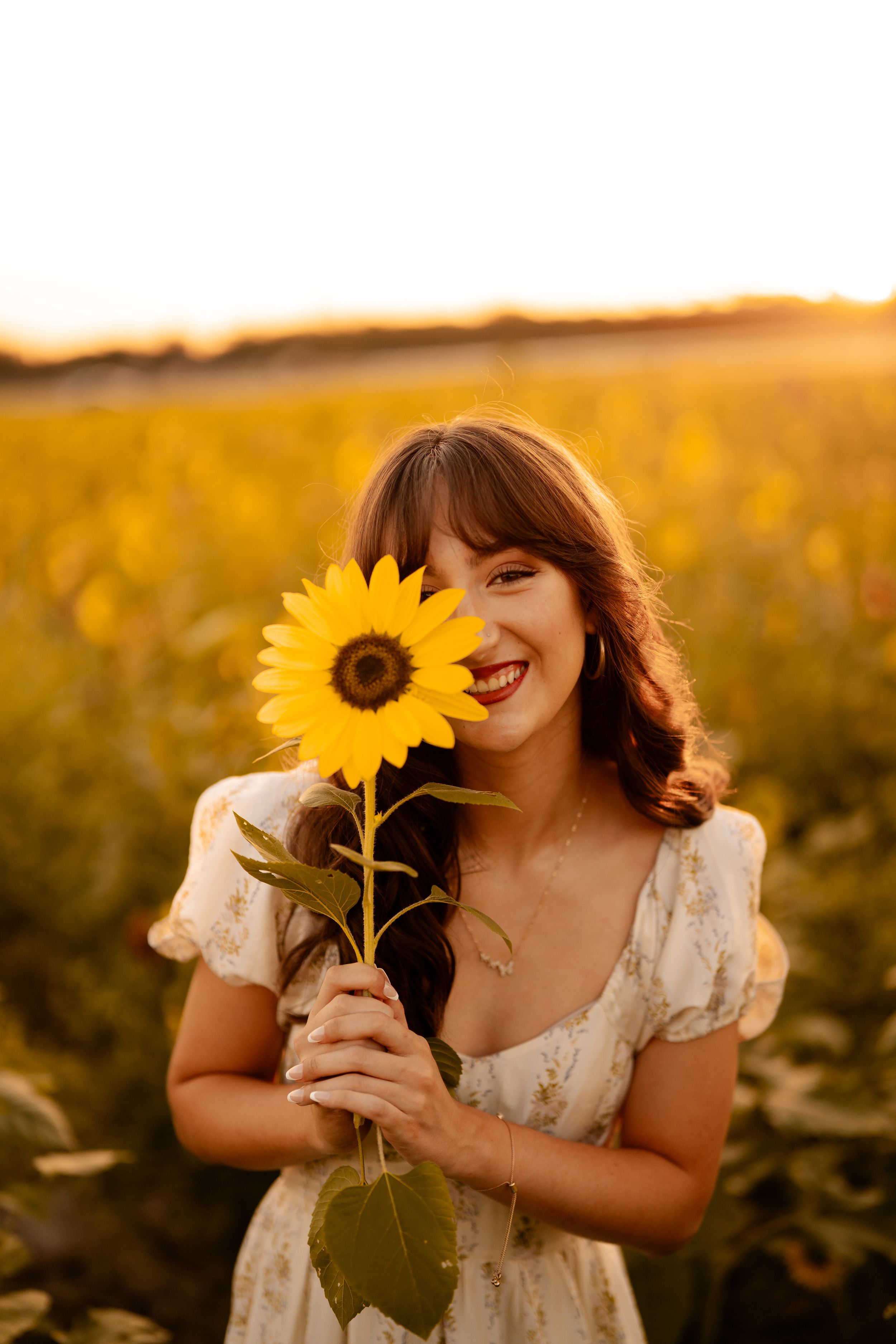 A bright sunflower is held up in a golden sunset field by someone wearing a white floral dress.
