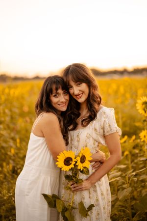 Two women embrace while holding sunflowers in a golden field at sunset.
