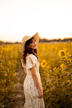 A woman in a flowing white dress and straw hat stands joyfully among golden sunflowers at sunset.