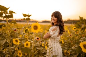 Young woman in white dress poses in golden sunflower field at sunset with warm lighting and dreamy atmosphere.