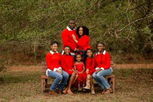Family wearing coordinating red sweaters poses together on wooden bench in outdoor nature setting.