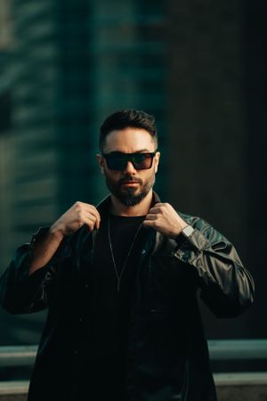 Person in sunglasses and black jacket adjusts collar against blurred urban background in moody lighting.