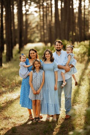 Family of five wearing coordinated blue and white outfits poses together in a sunlit forest setting.