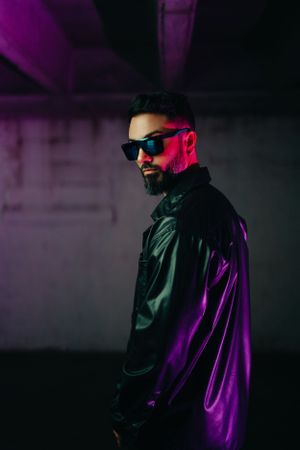 Cool portrait in sunglasses and black jacket under dramatic purple lighting against dark background.