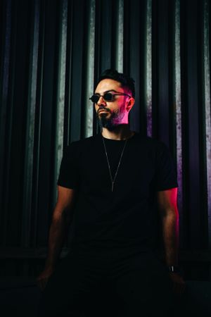 Person wearing sunglasses and black t-shirt stands against industrial backdrop with dramatic neon lighting.