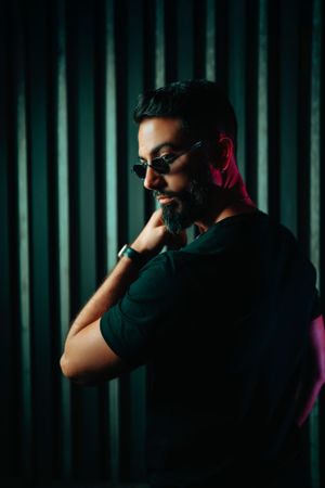 Dramatic portrait of person with beard and sunglasses posing against striped background with moody lighting and neon accents.