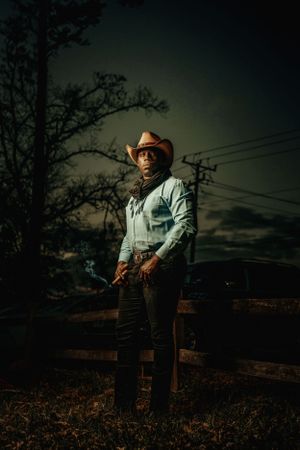 Dramatic nighttime portrait of a cowboy wearing a brown hat and denim shirt standing against a moody rural backdrop.