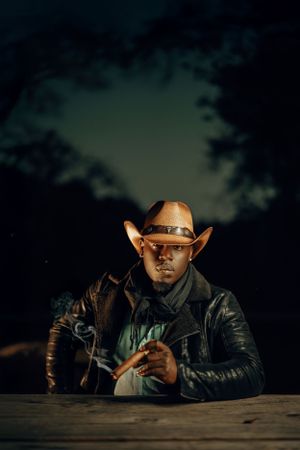 A person in a cowboy hat and leather jacket sits at a wooden table during nighttime under a moody sky.