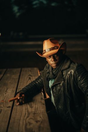 A dramatic portrait in low light shows someone in a brown cowboy hat and leather jacket pointing on wooden floorboards.