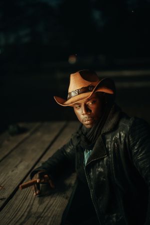 A person wearing a brown cowboy hat and black leather jacket sits thoughtfully on a wooden deck in moody lighting.