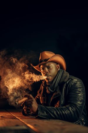 A figure in a brown cowboy hat and leather jacket exhales smoke while seated at a wooden table in dramatic lighting.