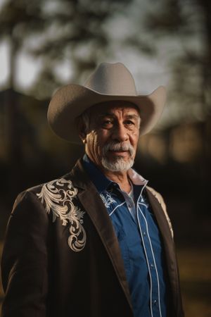A person in a brown embroidered western jacket and cowboy hat stands outdoors in warm lighting.
