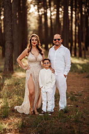 A family dressed elegantly poses in a pine forest during golden hour, with warm sunlight filtering through the trees.