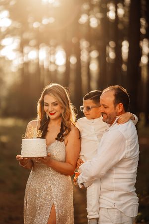 A family celebrates with a white cake in a sunlit forest during golden hour at sunset.