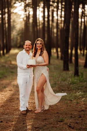 A couple poses together in a romantic forest setting at sunset wearing elegant white attire for their wedding photoshoot.