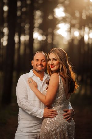 Couple embraces in a forest at golden hour with bokeh lighting and formal attire.