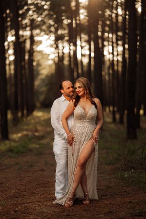 A couple embraces in a sunlit forest path wearing elegant white and sparkly formal attire.