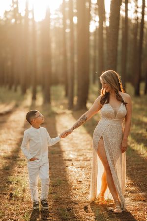 Mother and child holding hands in sunlit forest wearing white outfits during golden hour photo session.