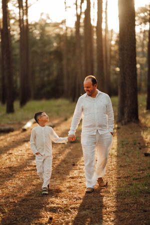 A parent and child walk hand in hand down a sunlit forest path wearing matching white outfits at golden hour.