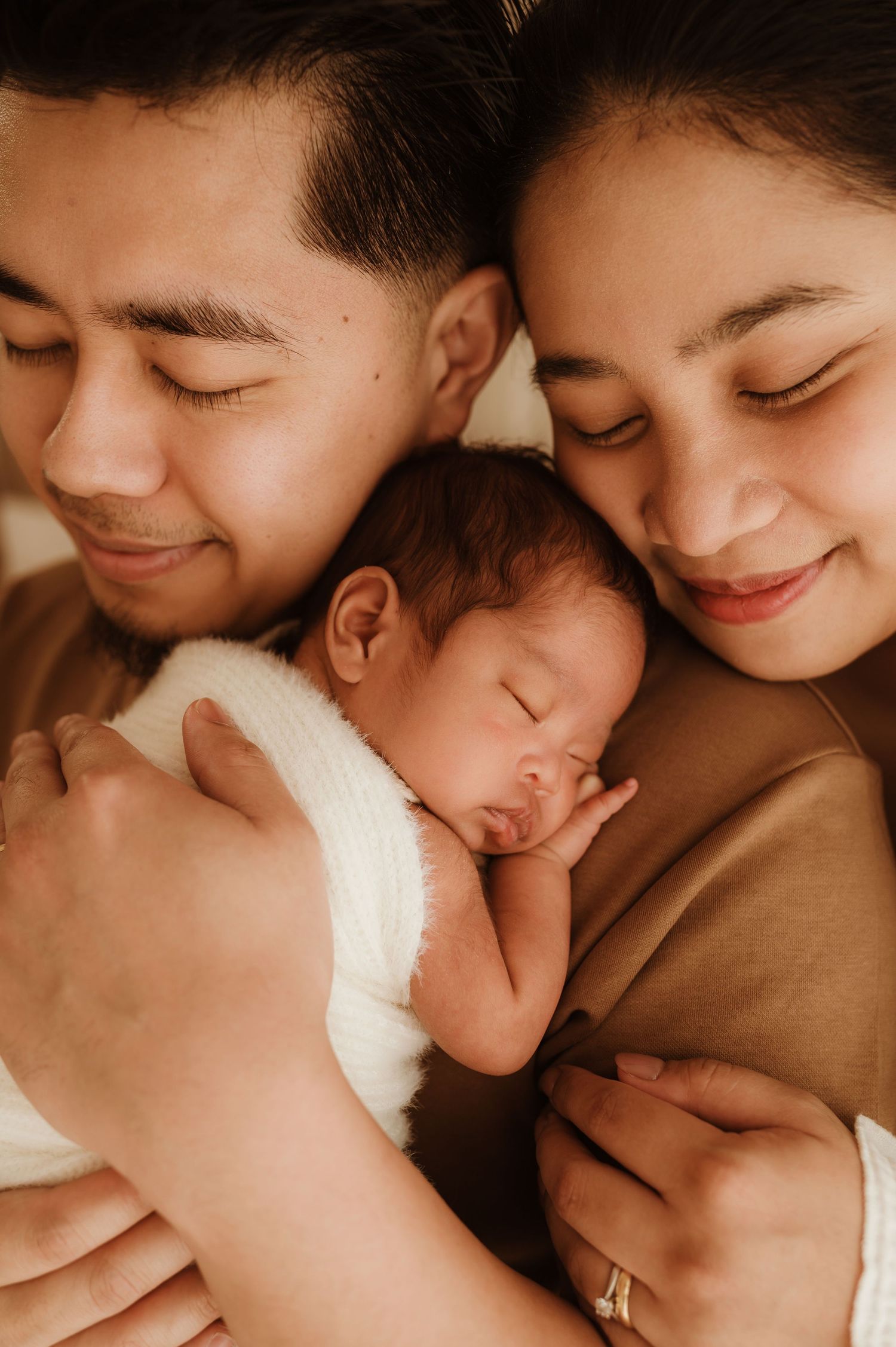 A small baby wrapped in a cozy white knit sweater lies on cream-colored fabric while a gentle hand rests on their head.