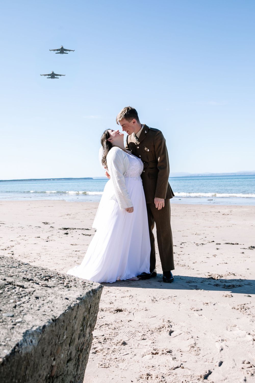 Two jets fly over a military wedding couple sharing a romantic moment on a sunny beach.