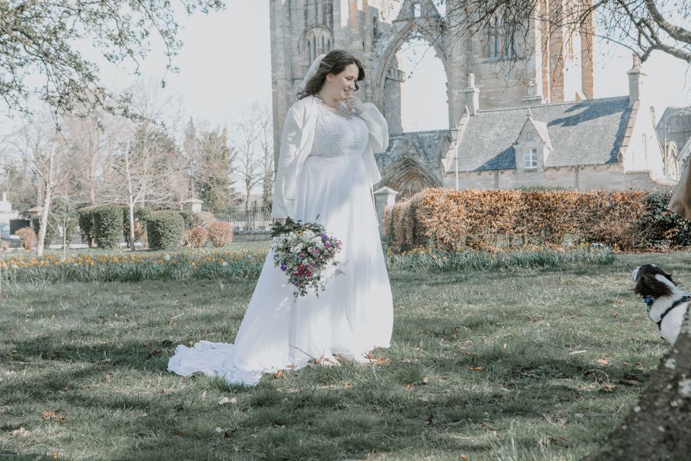 A person in an elegant white wedding dress stands on grass holding a bouquet near a gothic church structure.