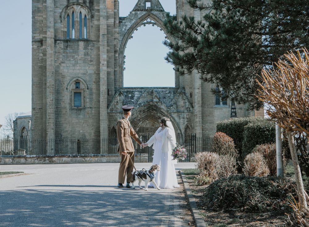 A couple in formal wedding attire takes their dog for a photo near a stone cathedral during a winter day.