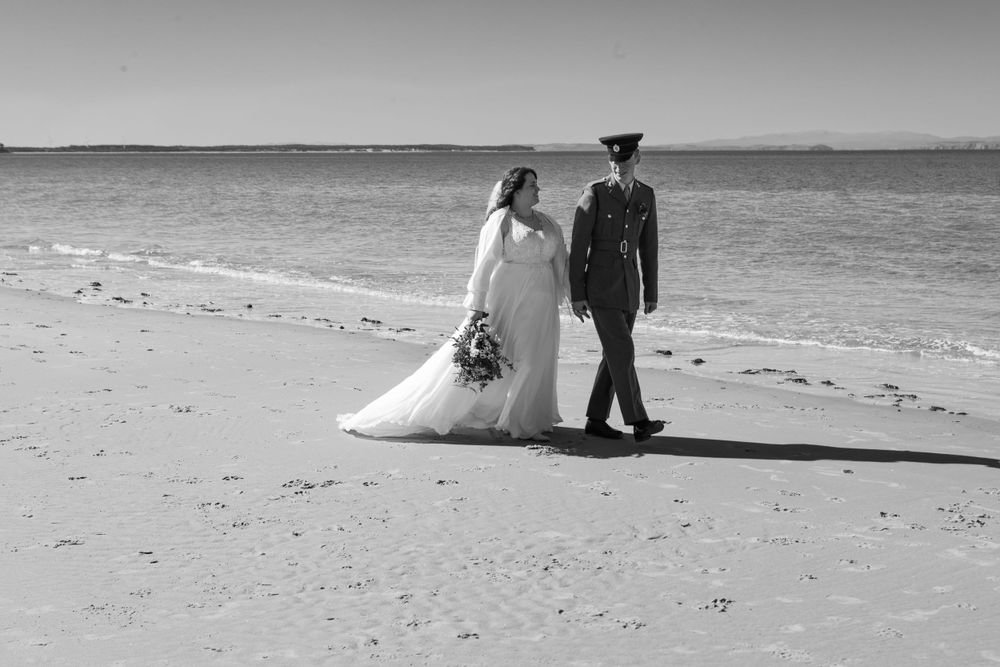 A newlywed couple in wedding attire walks along a sandy beach shore in black and white.