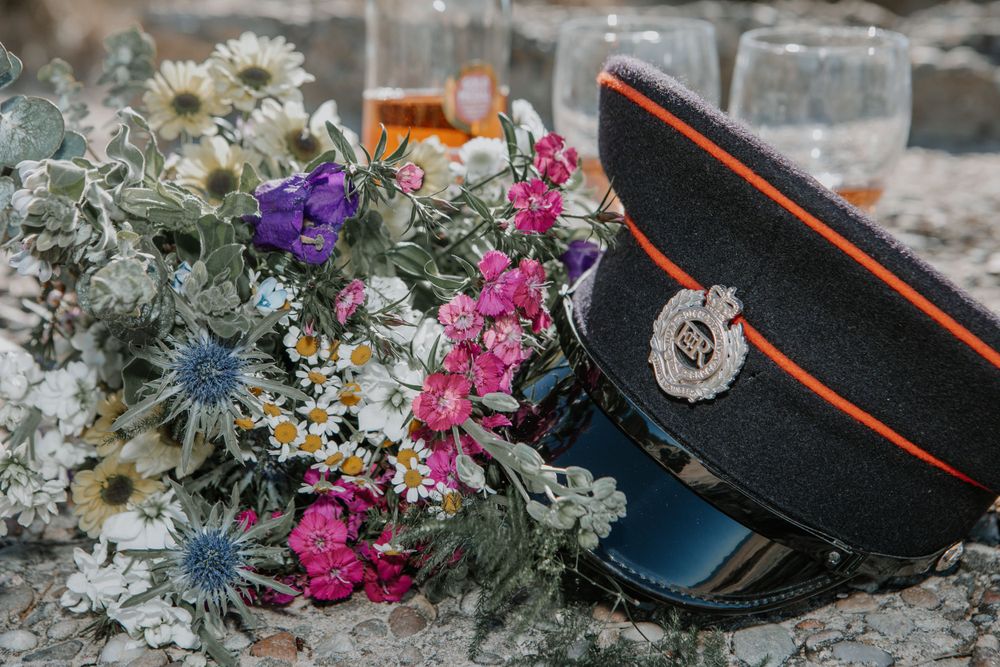 Military cap with orange trim sits beside colorful wildflower bouquet featuring daisies, thistle and pink blooms.